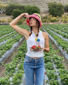 a woman standing in a strawberry field holding a plate with strawberries on it and wearing a pink hat