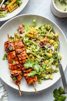 two plates with chicken, lettuce and corn on skewers next to salad