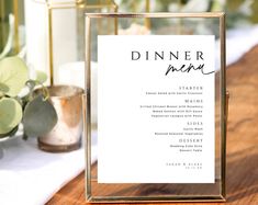 a table setting with a menu and flowers on the side, in front of a gold frame