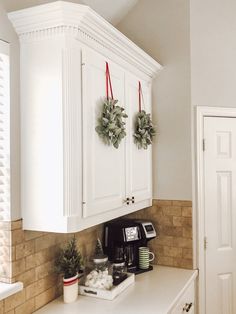 two wreaths hanging on the wall above a coffee maker in a kitchen with white cabinets