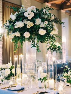 white flowers in a tall glass vase on top of a table with candles and place settings