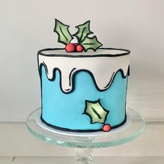 a decorated blue and white cake with holly decorations on it's top, sitting on a clear pedestal