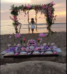 a table set up on the beach with flowers and plates in front of an arch