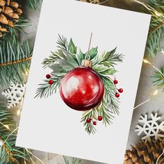 a watercolor christmas ornament on a card surrounded by pine cones and evergreen leaves