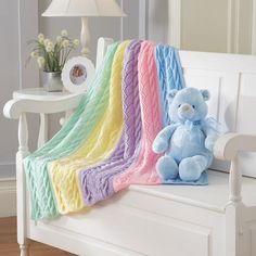 a blue teddy bear sitting on top of a white bench next to a rainbow blanket