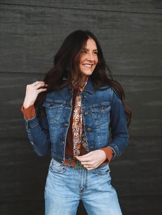 A woman with long dark brown hair standing in front of a wood wall wearing western fashion pieces great for spring including a medium wash jean jacket, wild rag, light wash blue jeans, and turquoise jewelry. Fall Button-up Denim Vest, Fall Denim Outerwear With Flap Pockets, Fall Denim Vest For Workwear With Buttons, Fall Medium Wash Denim Vest For Workwear, Button-up Denim Vest With Pockets For Fall, Medium Wash Denim Vest For Fall Workwear, Fall Denim Blue Vest With Buttons, Fall Denim Blue Jacket With Flap Pockets, Fall Denim Blue Denim Jacket With Flap Pockets