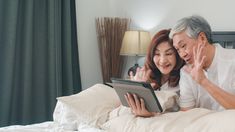 an older man and woman laying in bed looking at a tablet