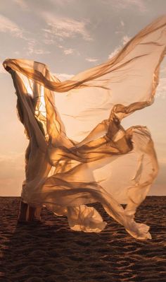 a woman standing on top of a sandy beach under a sheer white cloth flying in the air