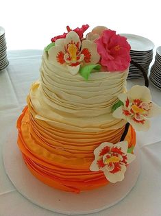 a multi - layered cake with flowers on top sits on a table in front of stacks of plates