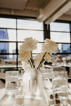 white flowers in a vase on a table with water and silverware next to large windows