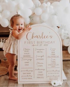 a baby girl standing next to a sign with balloons in the background that says, charles's first birthday