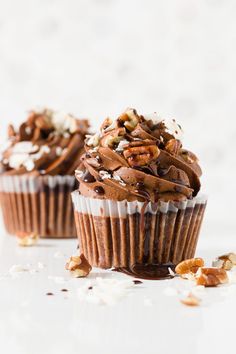 two cupcakes with chocolate frosting and nuts on top, sitting on a white surface