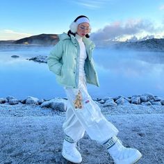 a woman in white pants and jacket walking on the ground near water with mountains in the background