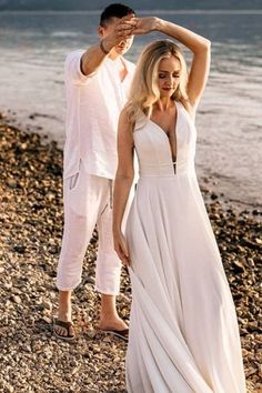 a woman in a white dress standing next to a man on the beach with his hands behind his head