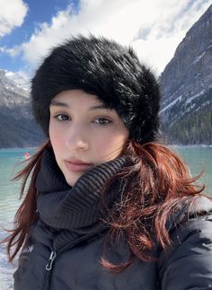 a young woman wearing a black hat and scarf standing in front of a mountain lake