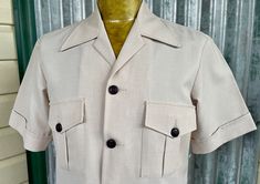 Show off your retro-style in this awesome 1970's vintage safari jacket. This funky jacket is a versatile cream colour and has an exaggerated 70's pointy collar, short cuffed sleeves and large black buttons for contrast. The front features four patch pockets with pointed flaps and decorative pleats, perfect for storing all adventure essentials. The back also has a large centre pleat and long single vent in keeping with the classic safari style.  Size Large  Measurements: Chest: 109 cm / 43 inches Shoulder: 46 cm / 18.25 inches Length: 73.5 cm / 29 inches Sleeve: 25 cm / 10 inches This item is one of a kind. BUYING VINTAGE:                                                                                                                                           While all care is taken to state Khaki Collared Outerwear, Vintage Short Sleeve Outerwear For Spring, Vintage Spring Outerwear With Short Sleeves, Vintage Short Sleeve Spring Outerwear, Summer Collared Outerwear With Flap Pockets, Vintage Outerwear With Pockets And Short Sleeves, Vintage Beige Outerwear With Snap Buttons, Cream Outerwear With Lapel Collar And Welt Pockets, Cream Outerwear With Lapel Collar