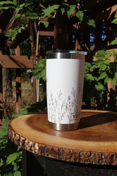 a white coffee cup sitting on top of a wooden table next to some trees and bushes