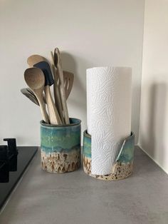 two cups with spoons and utensils in them sitting on a counter next to a roll of toilet paper