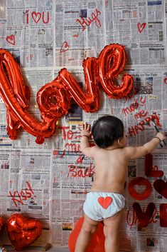 a baby in diapers reaching up to the wall with red balloons that spell out love