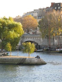 two people sitting on a bench near the water in front of a bridge and buildings