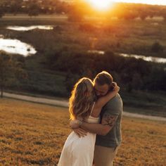 a man and woman embracing each other in an open field with the sun setting behind them