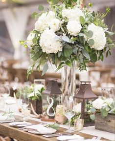 a tall vase filled with white flowers sitting on top of a table covered in greenery