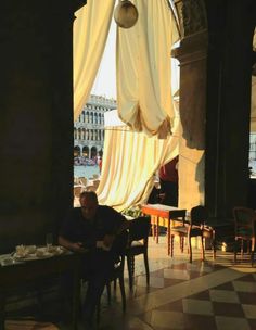 a man sitting at a table in front of a window with curtains hanging from it