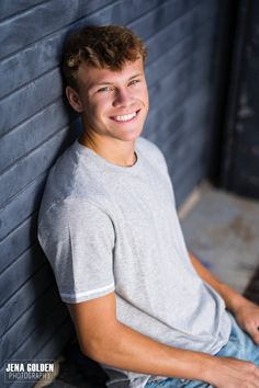 a young man sitting against a brick wall with his hand on his hip and smiling at the camera