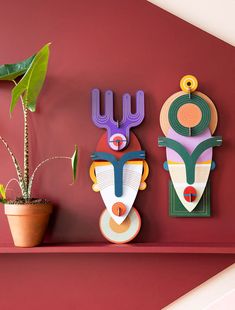 two wooden masks on a red wall next to a potted plant in a vase