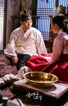 a man and woman sitting on a bed next to each other in front of a bowl