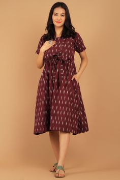 a woman in a maroon dress standing on a tan background with her hands behind her back