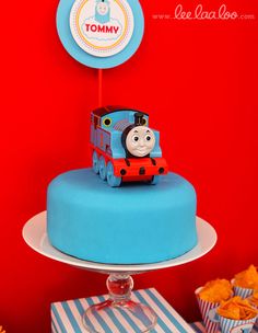 a thomas the train birthday cake with cupcakes around it and a red wall in the background