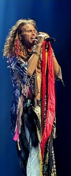 a man with long hair and feathers on stage holding a microphone in his right hand