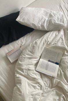 an open book is laying on top of a bed with white sheets and black pillows