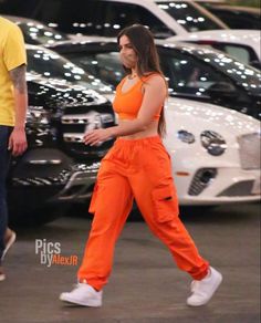 a woman in an orange jumpsuit is walking past parked cars