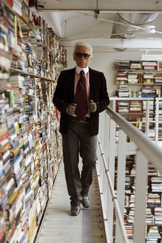 an older man in a suit and tie walking down a long hallway with stacks of books on the wall behind him