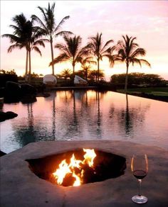 an outdoor fire pit with two glasses of wine on the table and palm trees in the background