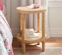 a wicker side table with books on it in front of a bed and window