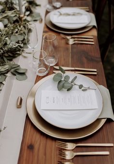 the table is set with white plates and silverware, greenery, and gold cutlery