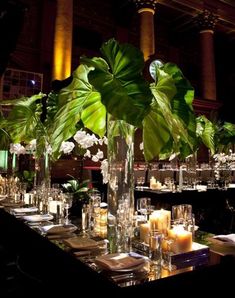 a long table is set with candles and vases filled with greenery, surrounded by tall pillars