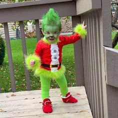 a young child dressed in green and red is standing on a porch with her arms out