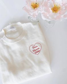 a white sweater with a red heart embroidered on the chest next to some pink flowers