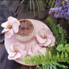 a pink plate topped with a cup filled with liquid and flowers next to purple plants