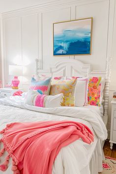 a white bed topped with lots of pillows next to a painting on the wall above it