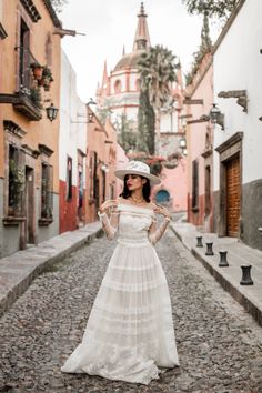 a woman in a white dress and hat standing on a cobblestone street