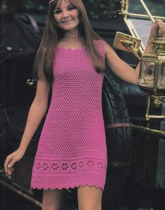 a woman in a pink crochet dress standing next to an old car and smiling