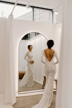 a woman standing in front of a mirror wearing a wedding dress with long sleeves and open back