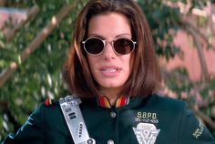 a woman wearing sunglasses and a green jacket is standing in front of a tree with her hand on the belt