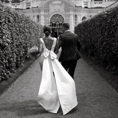 a bride and groom walking down a path in front of a building with hedges on either side