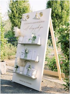 a white sign with flowers and candles on it in front of some trees, bushes and other greenery